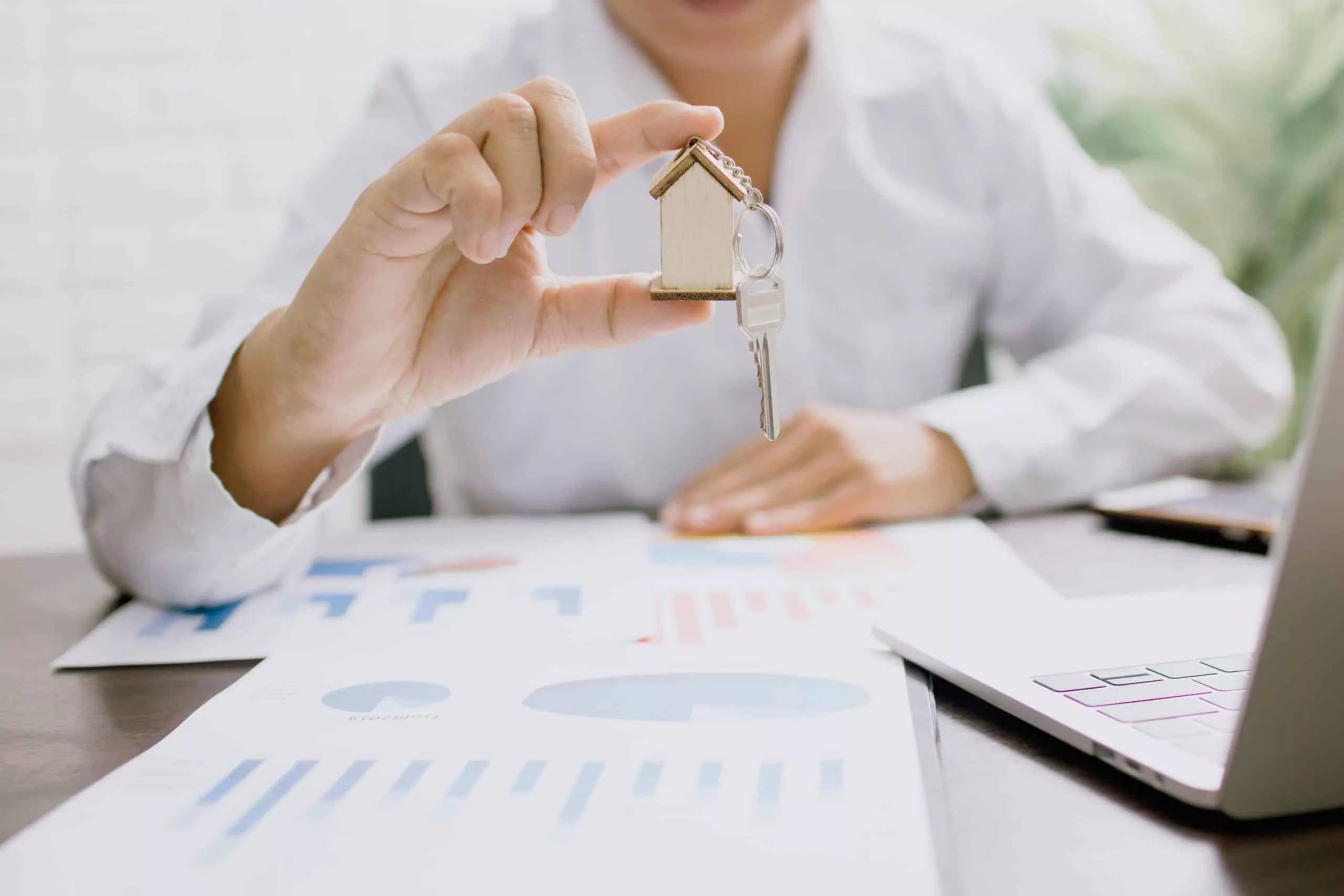 Person holding a house key fob above printed cost charts and a laptop