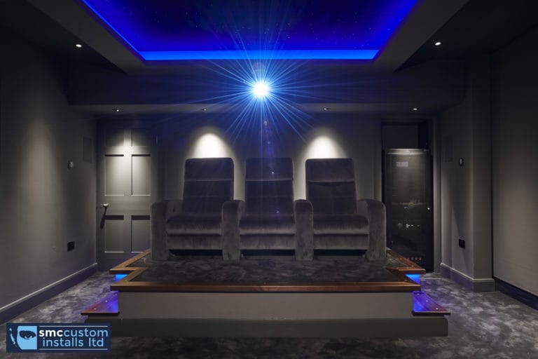 Three velvet recliners on raised platform beneath blue LED ceiling in dedicated home cinema