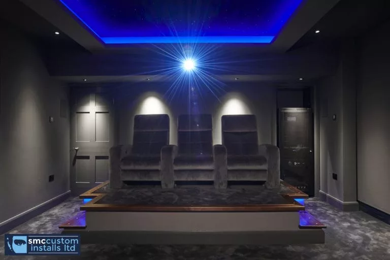 Three velvet recliners on raised platform beneath blue LED ceiling in dedicated home cinema