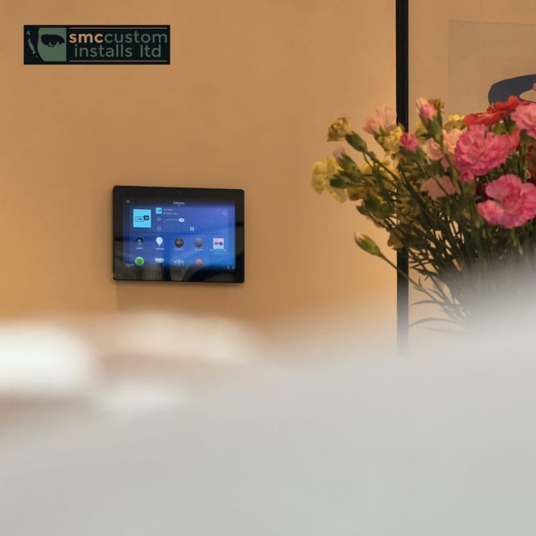 Wall-mounted smart home touchscreen control panel beside a vase of fresh flowers