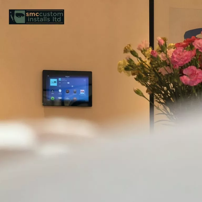 Wall-mounted smart home touchscreen control panel beside a vase of fresh flowers
