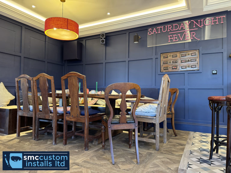 Dining corner of a blue-panelled bar room with wall-mounted speakers and neon sign