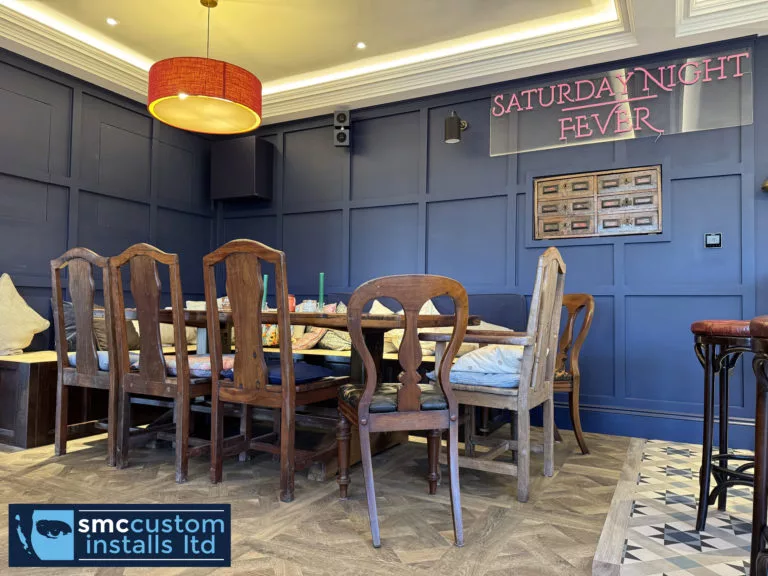 Dining corner of a blue-panelled bar room with wall-mounted speakers and neon sign