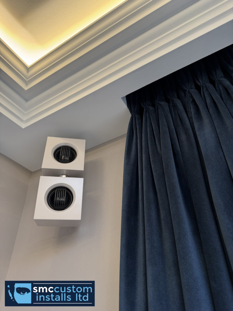 Pair of wall-mounted surround speakers beside curtains beneath LED-lit coving