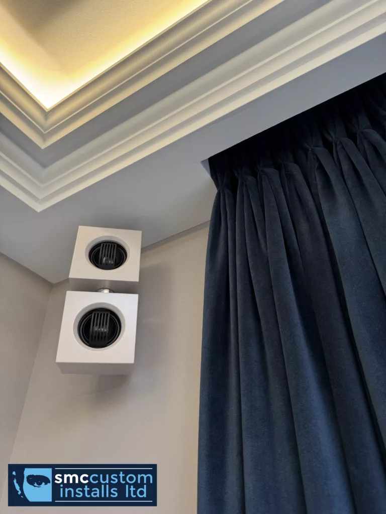 Pair of wall-mounted surround speakers beside curtains beneath LED-lit coving