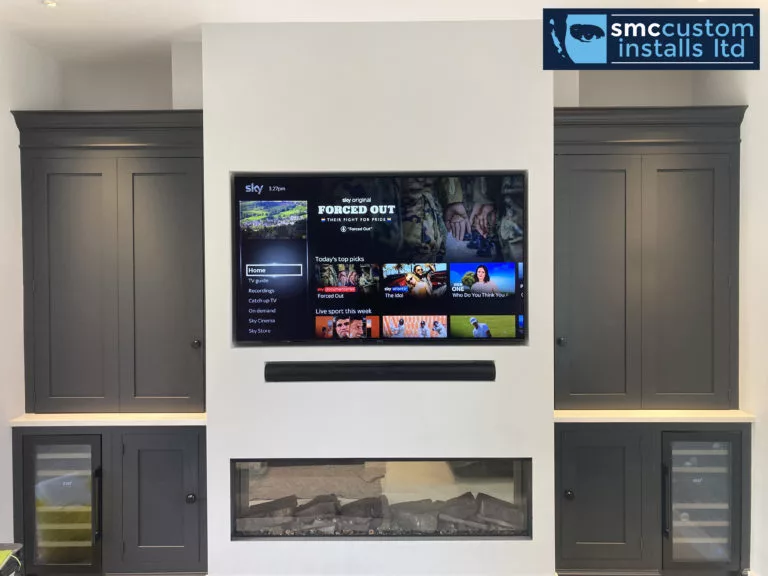 Wall-mounted TV and soundbar above an inset fire, flanked by cabinets and drinks fridges