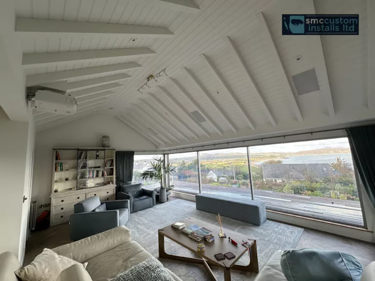 Ceiling-mounted projector and in-ceiling speakers in a vaulted living room with panoramic glazing