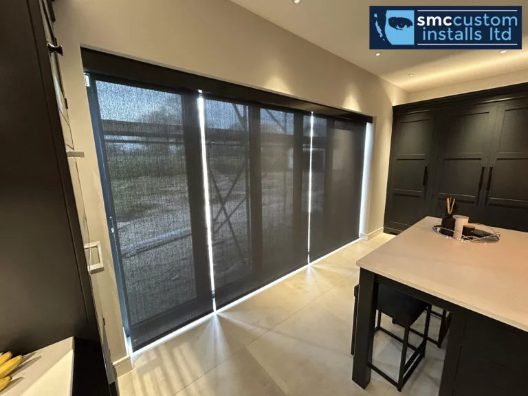 Motorised roller blinds lowered across full-height glazed doors in a dark kitchen